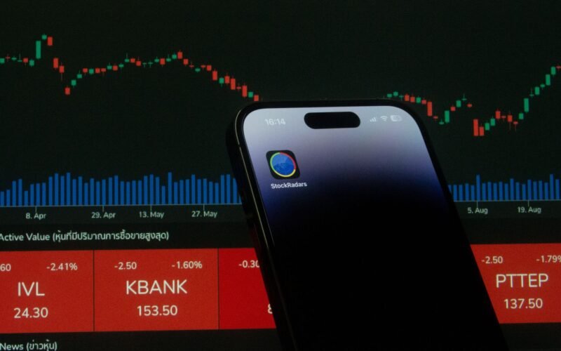 A mobile phone displaying a trading app in front of a financial market chart screen.