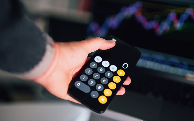 Close-up of a hand holding a smartphone calculator with financial charts on a screen in the background.