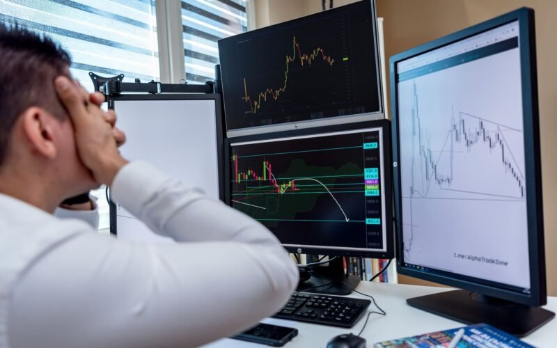 Frustrated man monitoring multiple trading graphs on computer screens in an office setting.