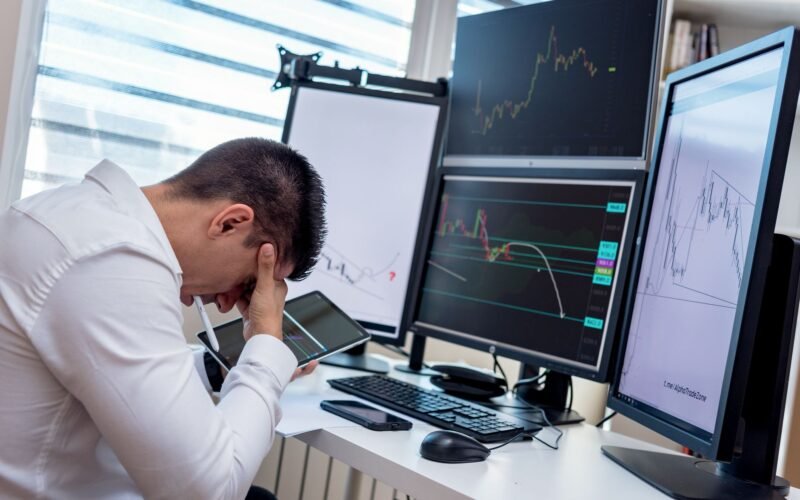 A stressed trader in an office setting analyzes market data on multiple monitors using a tablet.