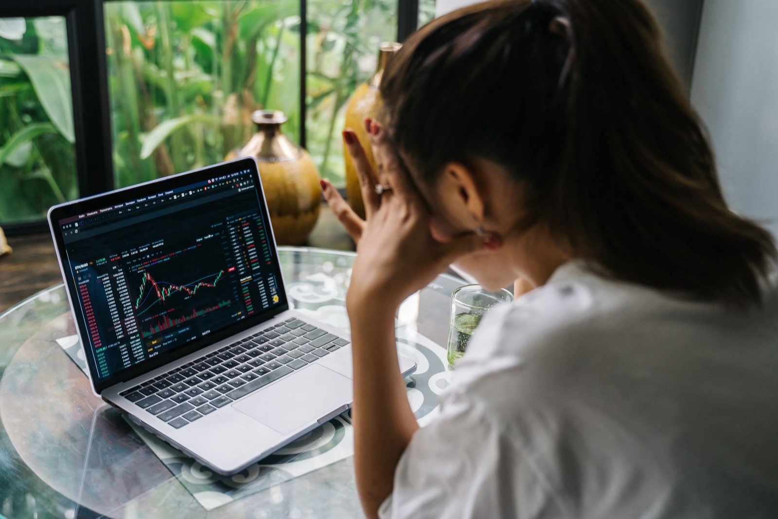 Woman analyzing cryptocurrency market charts on a laptop, looking stressed in a cozy indoor setting.