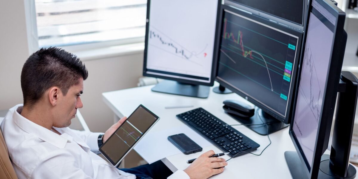 Business professional analyzing financial charts on monitors and tablet in modern office workspace.