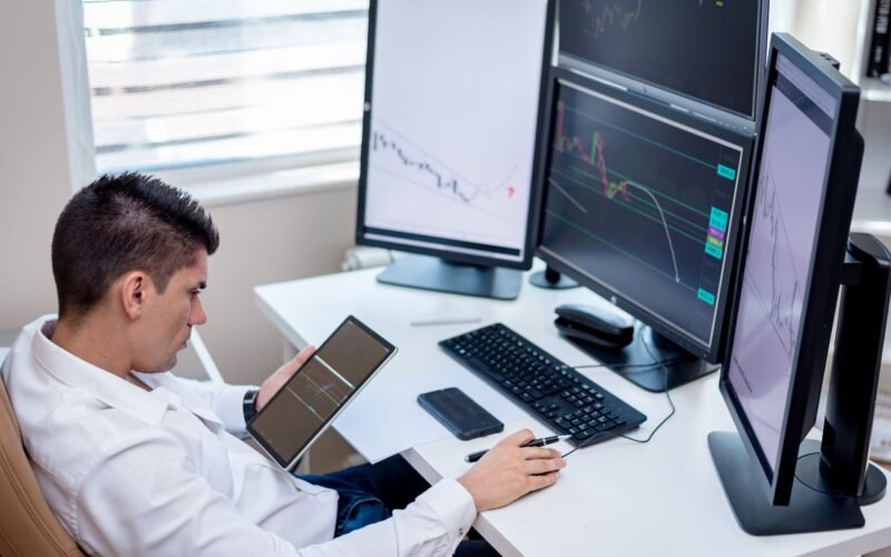 Business professional analyzing financial charts on monitors and tablet in modern office workspace.
