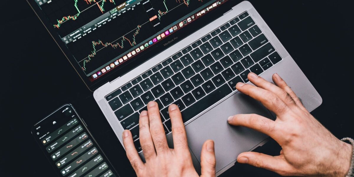Hands typing on a laptop displaying stock market charts and a smartphone with financial apps.