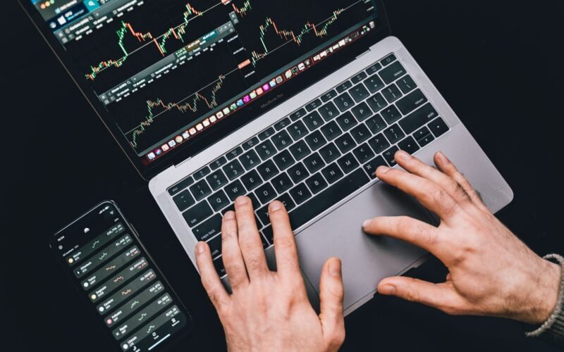 Hands typing on a laptop displaying stock market charts and a smartphone with financial apps.