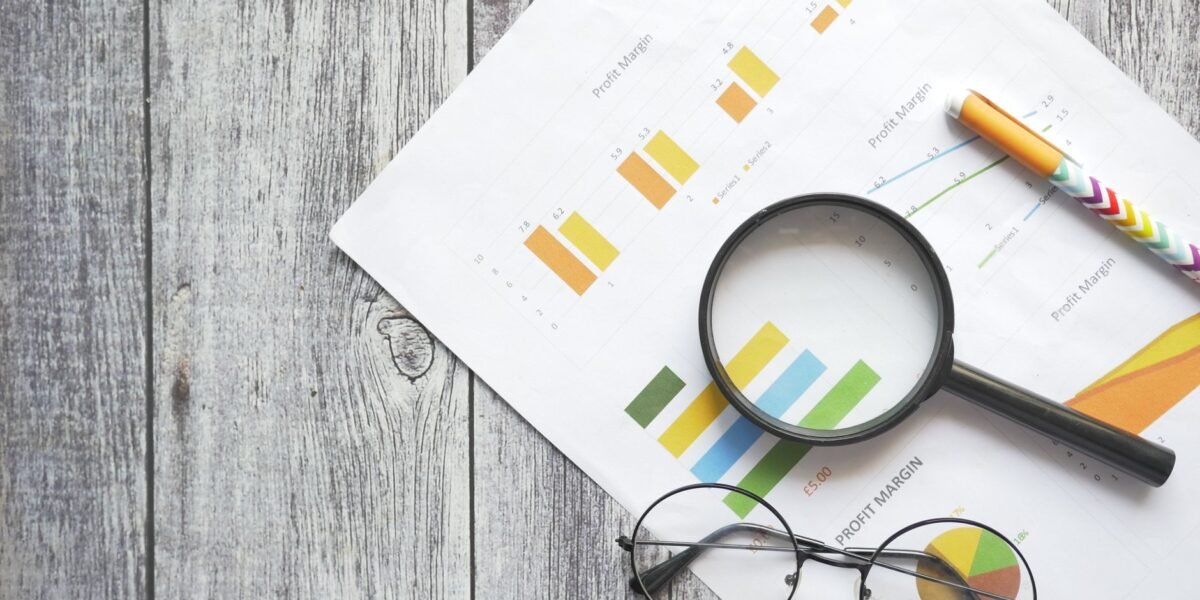Overhead view of financial charts, a magnifying glass, ballpen, and eyeglasses on a wooden surface.