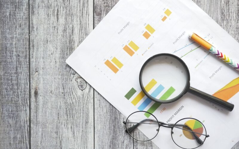 Overhead view of financial charts, a magnifying glass, ballpen, and eyeglasses on a wooden surface.