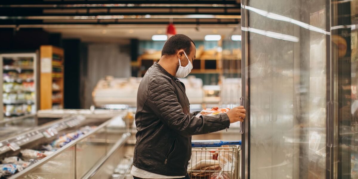 A man wearing a mask shops in a supermarket, selecting items from a refrigerated section.