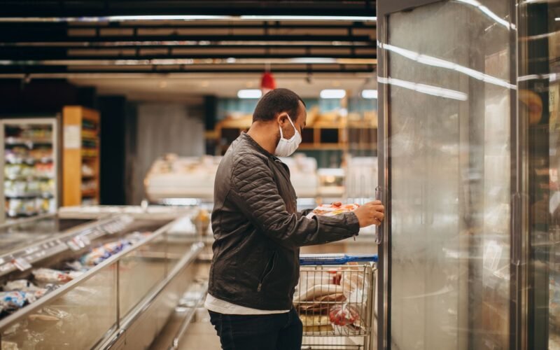 A man wearing a mask shops in a supermarket, selecting items from a refrigerated section.