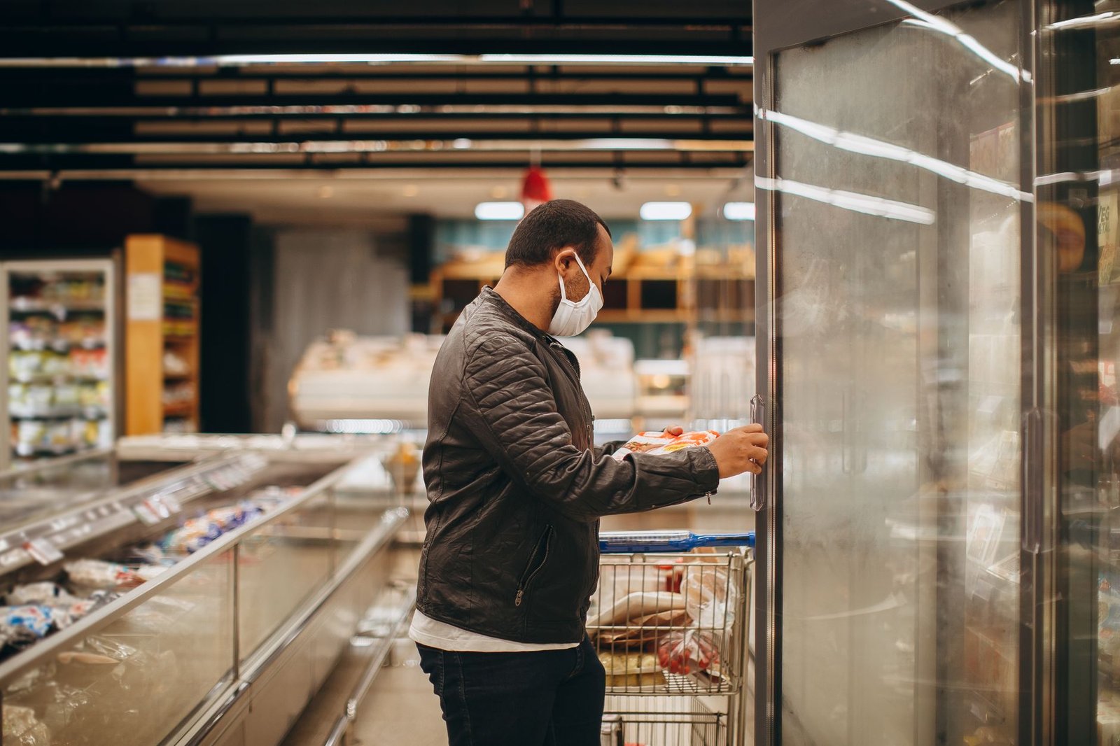 A man wearing a mask shops in a supermarket, selecting items from a refrigerated section.