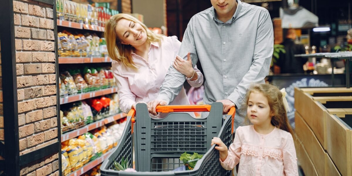 A family of three shopping in a supermarket, pushing a cart full of groceries.
