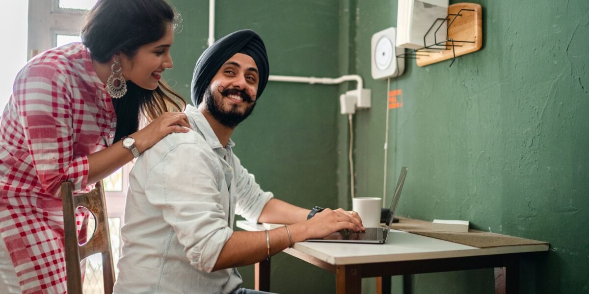 Smiling young Indian woman in casual clothes caring for positive husband working on laptop from home