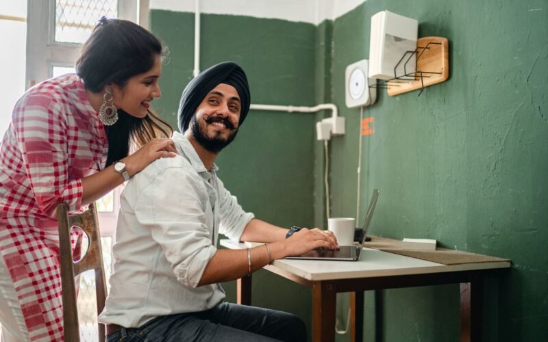 Smiling young Indian woman in casual clothes caring for positive husband working on laptop from home