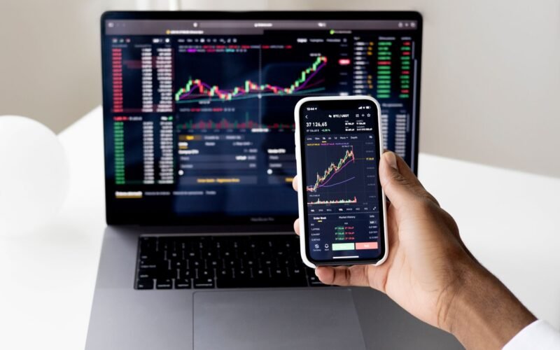 Person checking stock market graphs on laptop and smartphone, focusing on financial data.