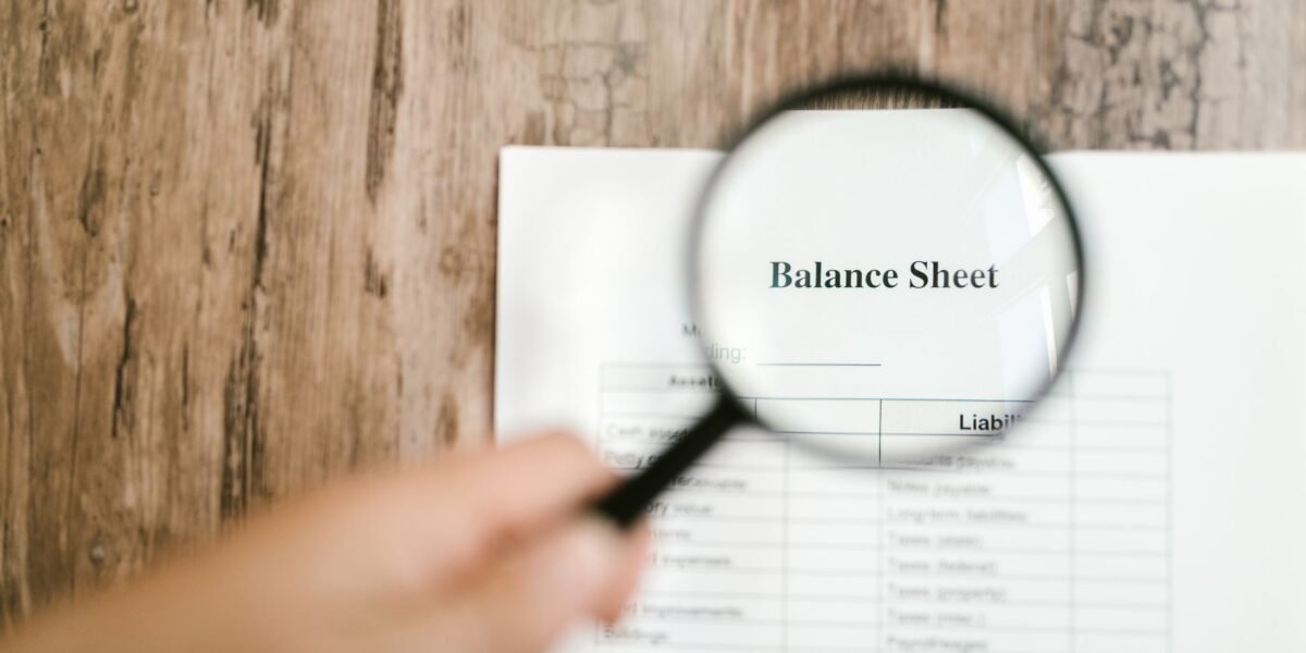 Close-up of a balance sheet document on wooden surface with a magnifying glass held by a hand.
