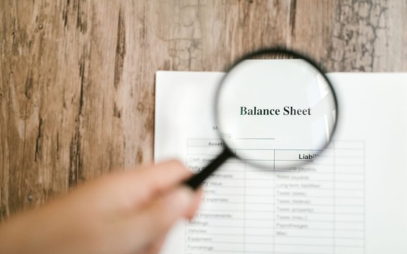 Close-up of a balance sheet document on wooden surface with a magnifying glass held by a hand.