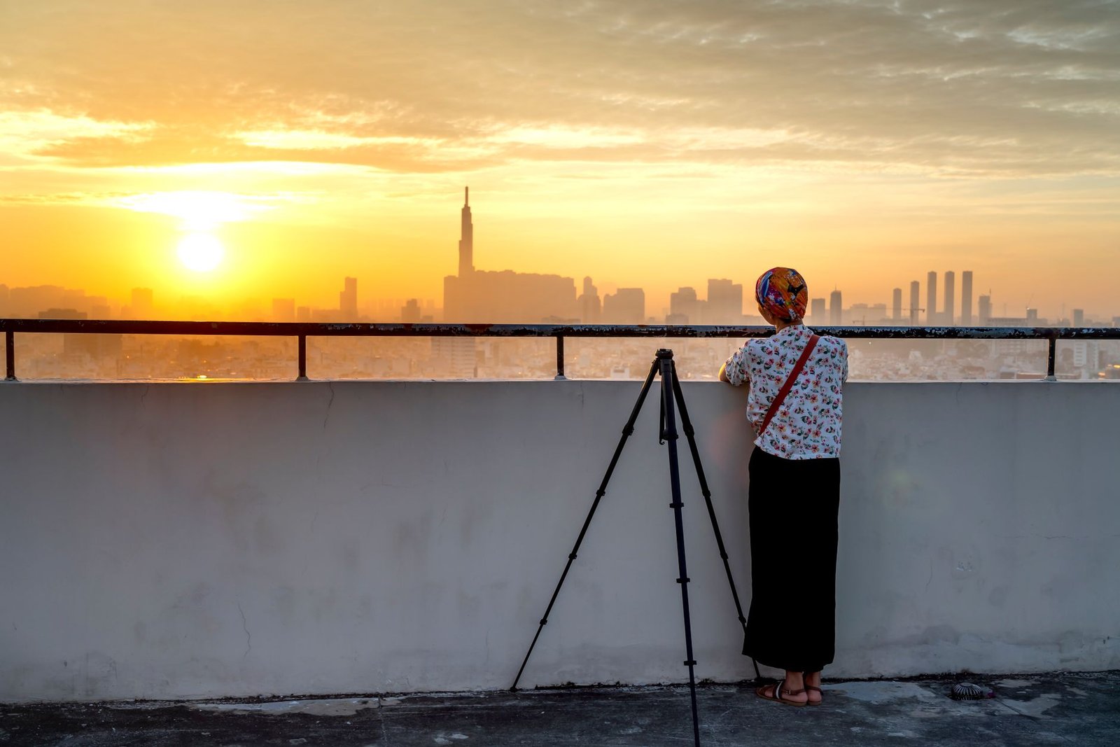 A woman with a tripod overlooking a cityscape during sunset from a rooftop.