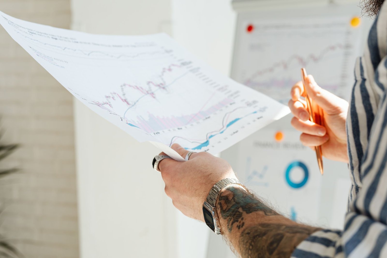 Close-up of a tattooed man's hands holding financial charts, with focus on data analysis and stock market trends.